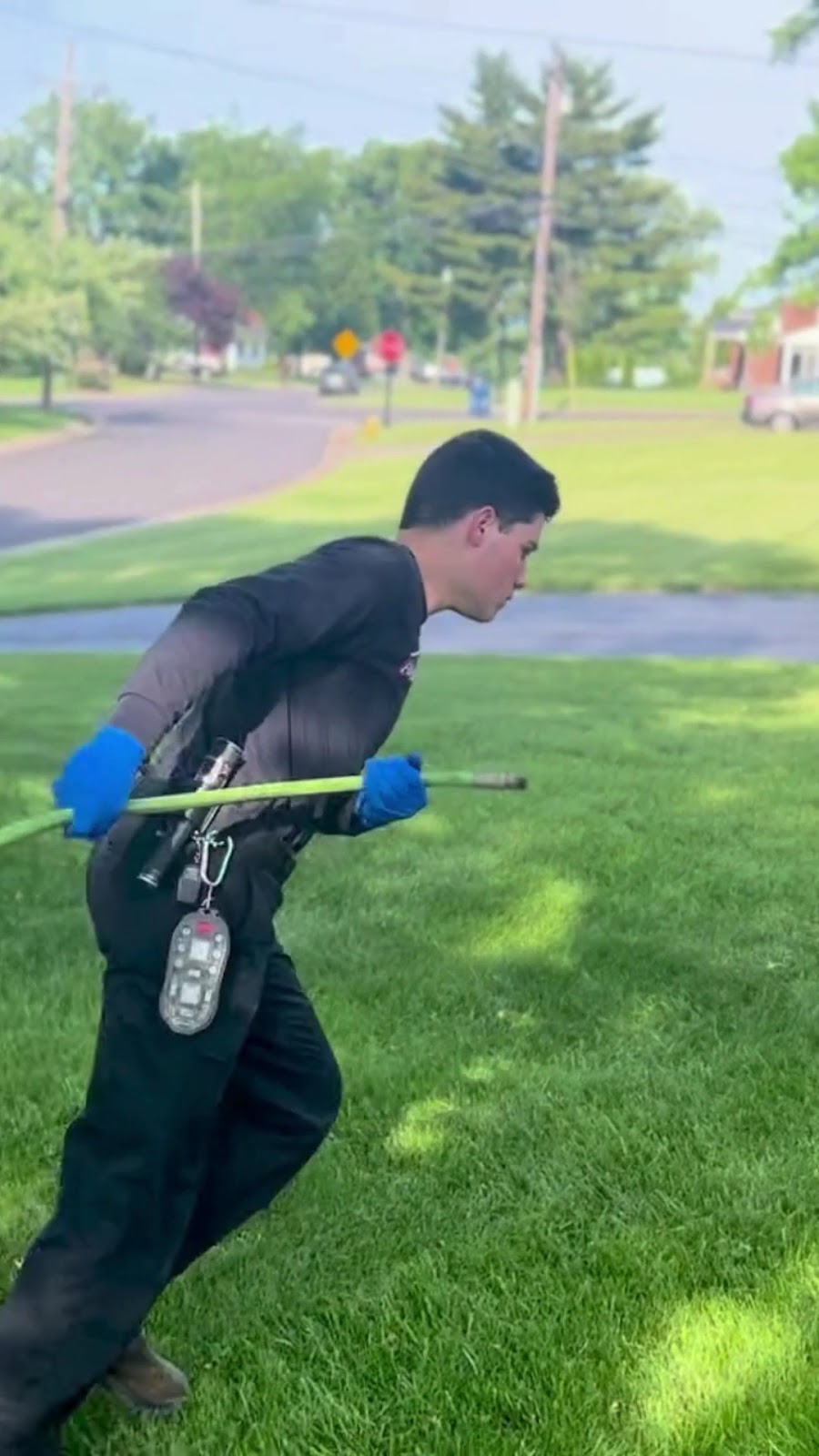 A technician wearing blue gloves and a work uniform runs across a green residential lawn holding a green hose.